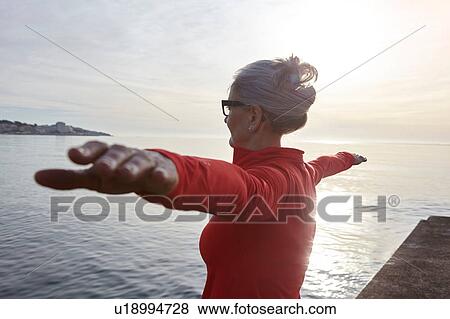 Stock Photo - Mature woman standing beside water, in yoga position, rear view. Fotosearch