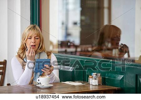 Mature woman using smartphone to apply lipstick at sidewalk cafe table View Large Photo Image Picture - Mature woman using smartphone to apply lipstick at sidewalk cafe table. Fotosearch