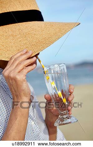 Stock Image - Mature woman wearing straw hat, beside beach, drinking cool drink through drinking straw. Fotosearch