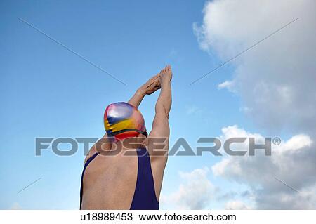 Stock Image - Mature woman, wearing swimming costume, arms raised, preparing to dive, rear view. Fotosearch
