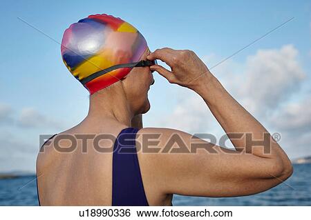 Mature woman wearing swimming costume and swimming hat, standing beside the sea, adjusting swimming goggles, rear view View Large Photo Image Stock Photograph - Mature woman wearing swimming costume and swimming hat, standing beside the sea, adjusting swimming goggles, rear view. Fotosearch