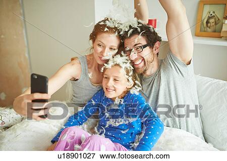 Mid adult couple and daughter covered in pillow fight feathers taking smartphone selfie in bed View Large Photo Image Stock Photo - Mid adult couple and daughter covered in pillow fight feathers taking smartphone selfie in bed. Fotosearch
