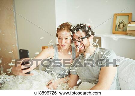 Stock Photograph - Mid adult couple covered in pillow fight feathers taking smartphone selfie in bed. Fotosearch