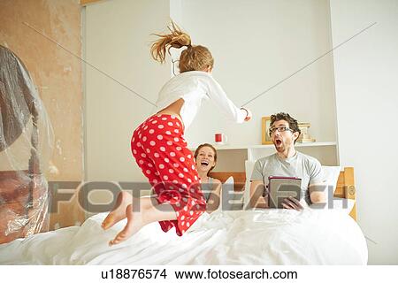 Picture - Mid adult couple having lie in whilst daughter jumps on bed. Fotosearch