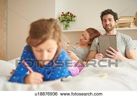 Stock Photograph - Mid adult couple having lie in whilst daughter draws on bed. Fotosearch