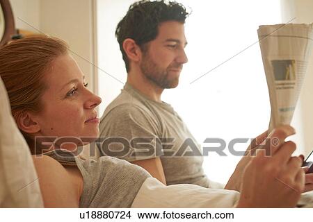 Picture - Mid adult couple reading broadsheet in bed. Fotosearch