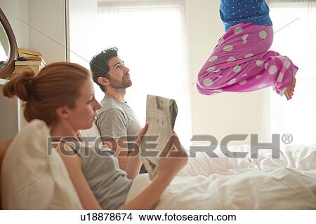 Picture - Mid adult couple reading broadsheet whilst daughter jumps on bed. Fotosearch