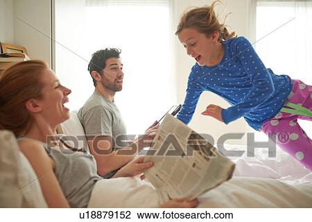Stock Image - Mid adult couple reading broadsheet whilst daughter leaps on bed. Fotosearch