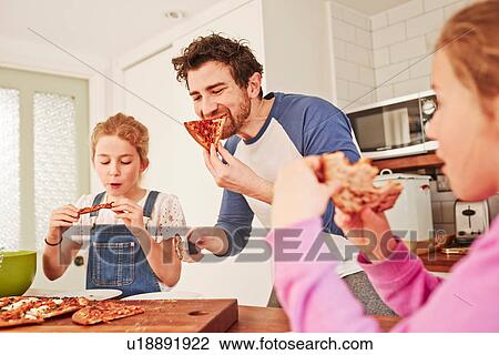 Stock Image - Mid adult man eating pizza with daughters at kitchen bench. Fotosearch