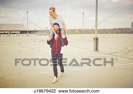 Stock Image - Mid adult man giving shoulder ride to girlfriend on rooftop parking lot. Fotosearch