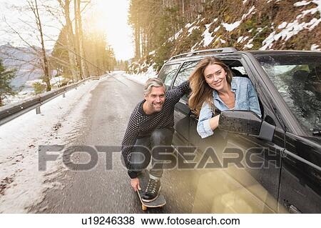Stock Photo - Mid adult man on skateboard, holding onto side of car, smiling. Fotosearch