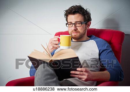 Stock Photo - Mid adult man reading book on armchair. Fotosearch