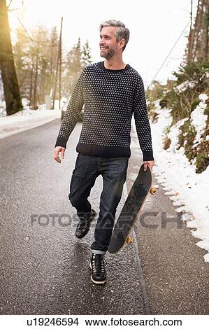 Picture - Mid adult man walking along road, carrying skateboard. Fotosearch