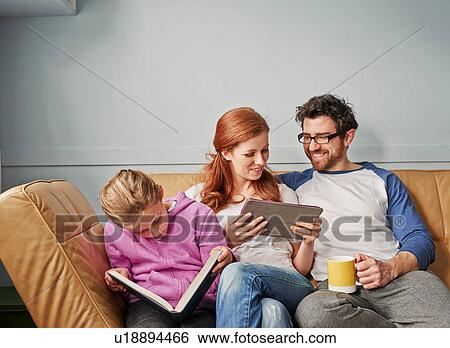 Stock Photograph - Mid adult parents and daughter on sofa on reading book and digital tablet. Fotosearch