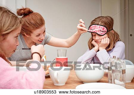Stock Image - Mid adult woman at breakfast table peeking under daughters sleep mask. Fotosearch
