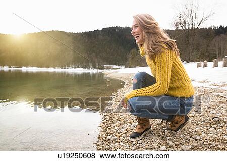 Stock Photo - Mid adult woman crouching beside lake, smiling, German Alps, Germany. Fotosearch