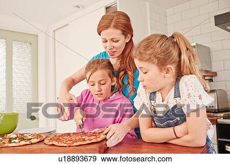 Stock Photo - Mid adult woman slicing pizza for daughters at kitchen bench. Fotosearch