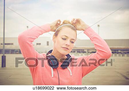 Stock Image - Mid adult woman wearing headphones tying hair on rooftop parking lot. Fotosearch