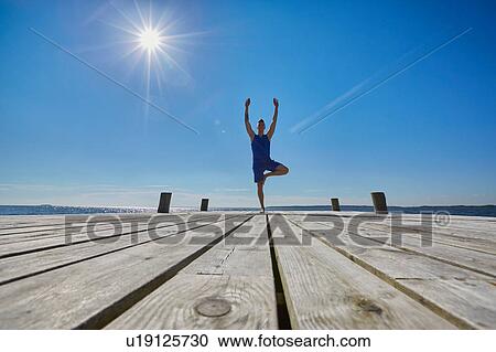 Stock Image - Mid distant view of man on pier, standing on one leg arms raised. Fotosearch
