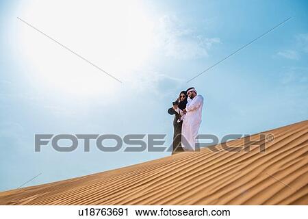 Stock Image - Middle eastern couple wearing traditional clothes taking smartphone selfie on desert dune, Dubai, United Arab Emirates. Fotosearch
