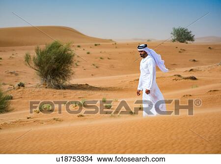 Picture - Middle eastern man wearing traditional clothes walking in desert, Dubai, United Arab Emirates. Fotosearch