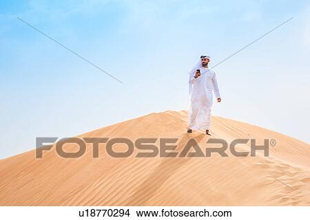Middle eastern man wearing traditional clothes using smartphone on desert dune, Dubai, United Arab Emirates View Large Photo Image Picture - Middle eastern man wearing traditional clothes using smartphone on desert dune, Dubai, United Arab Emirates. Fotosearch