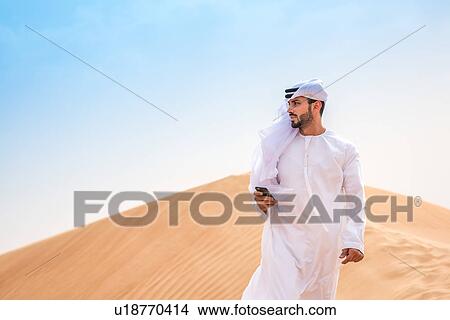 Middle eastern man wearing traditional clothes using smartphone on desert dune, Dubai, United Arab Emirates View Large Photo Image Picture - Middle eastern man wearing traditional clothes using smartphone on desert dune, Dubai, United Arab Emirates. Fotosearch