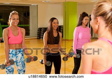 Stock Image - Mirror image of women in gym using dumbbells. Fotosearch