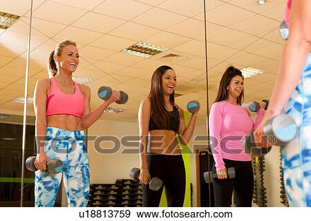 Stock Photo - Mirror image of women in gym using dumbbells. Fotosearch