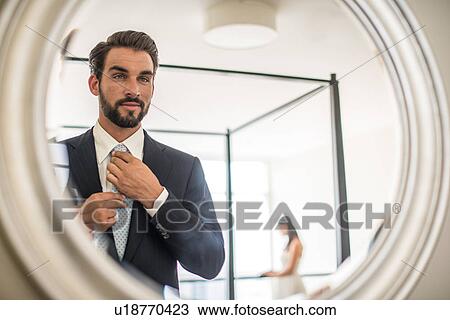 Stock Image - Mirror reflection of young businessman adjusting shirt and tie in hotel room, Dubai, United Arab Emirates. Fotosearch
