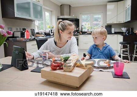 Stock Photo - Mother and son sitting at table eating meal together, son looking at digital tablet. Fotosearch