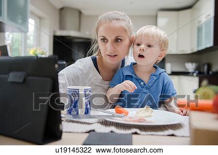 Stock Image - Mother and son sitting at table, looking at digital tablet. Fotosearch