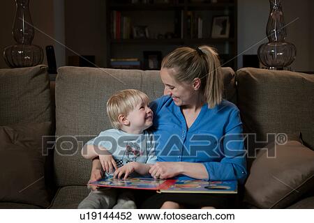 Stock Image - Mother and son sitting on sofa, reading book together. Fotosearch