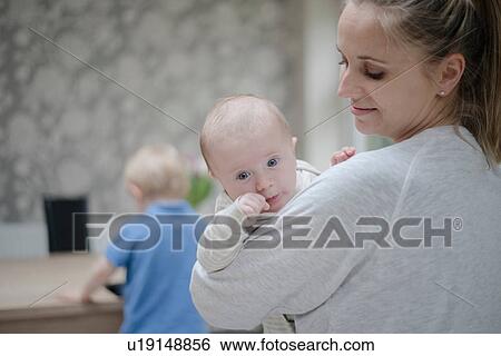 Stock Photograph - Mother holding baby boy, older son standing by table. Fotosearch