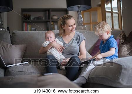 Stock Photograph - Mother sitting in living room with son and baby boy, son holding digital tablet. Fotosearch