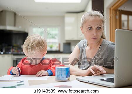 Stock Photo - Mother using laptop while son draws on paper beside her. Fotosearch