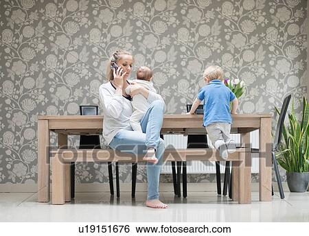 Mother using smartphone whilst holding baby boy, older son sitting beside her View Large Photo Image Stock Photograph - Mother using smartphone whilst holding baby boy, older son sitting beside her. Fotosearch