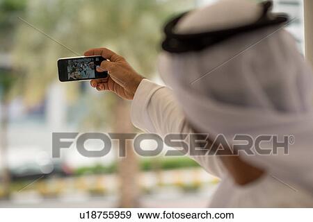Stock Photo - Over shoulder of middle eastern man taking smartphone selfie with friends at cafe, Dubai, United Arab Emirates. Fotosearch