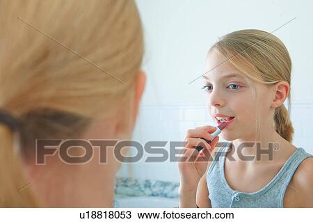 Stock Image - Over the shoulder mirror image of girl applying lipstick. Fotosearch