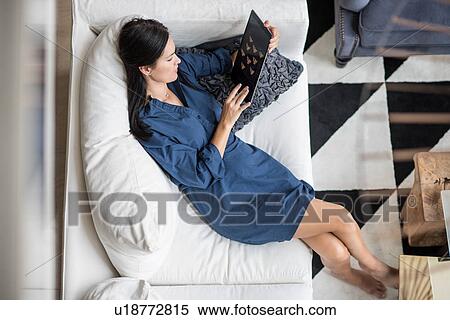 Stock Photography - Overhead view of woman reclining on hotel room sofa using digital tablet, Dubai, United Arab Emirates. Fotosearch