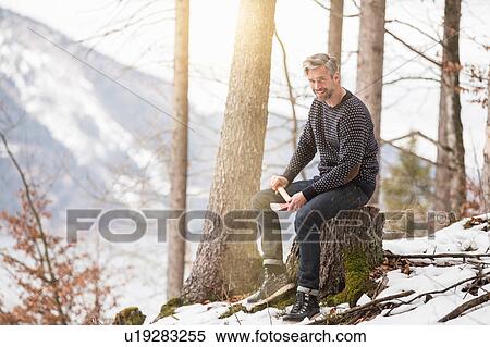 Stock Photography - Portrait of mid adult man sitting on tree stump, holding axe, German Alps, Germany. Fotosearch