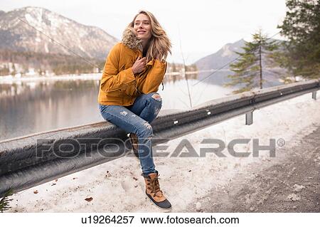 Stock Photo - Portrait of mid adult woman sitting on railing by side of road, German Alps, Germany. Fotosearch
