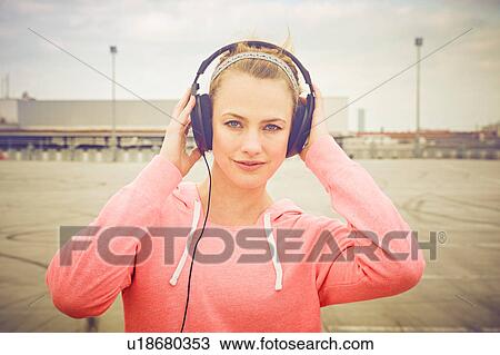 Stock Image - Portrait of woman with hands on headphones listening to music on rooftop parking lot. Fotosearch
