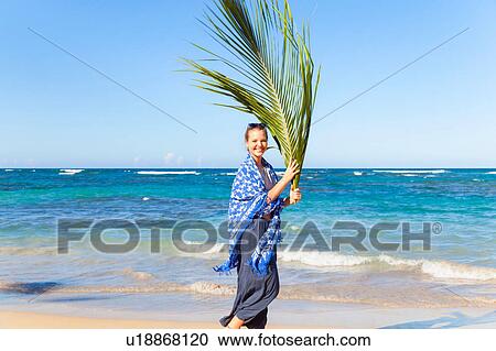 Stock Image - Portrait of young woman carrying palm leaf on beach, Dominican Republic, The Caribbean. Fotosearch