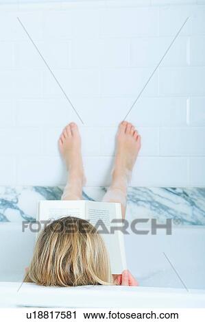 Stock Image - Rear view of blond haired girl reading a book in bath with feet up. Fotosearch