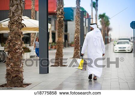 Rear view of man wearing dishdasha walking along street carrying shopping bags, Dubai, United Arab Emirates View Large Photo Image Stock Photo - Rear view of man wearing dishdasha walking along street carrying shopping bags, Dubai, United Arab Emirates. Fotosearch