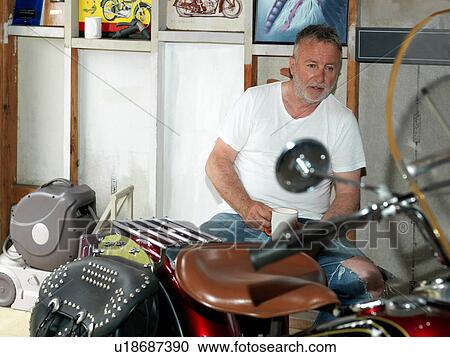 Stock Image - Senior man sitting with a coffee staring at motorcycle in garage. Fotosearch