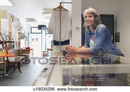 Stock Photo - Shop assistant leaning against glass counter looking away smiling. Fotosearch
