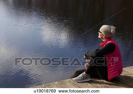 Stock Photo - Side view of woman sitting on wooden pier looking away. Fotosearch