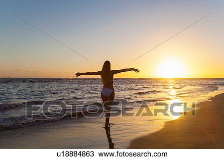 Stock Image - Silhouetted young woman with arms open on beach at sunset, Dominican Republic, The Caribbean. Fotosearch